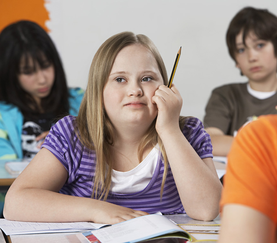 Teenage girl seated at desk in classroom, rests hand on face while holding a pencil between her fingers