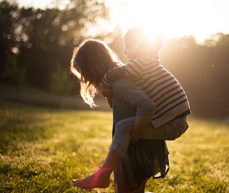Woman carrying young boy piggyback standing in field of grass. Sun shining in background.