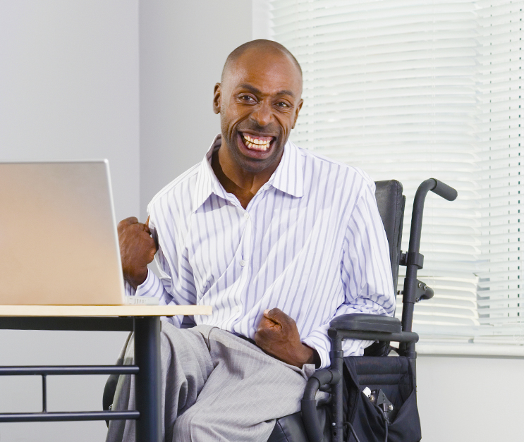 Middle-aged bald man with developmental disability sits in wheelchair in front of laptop and smiles at the camera