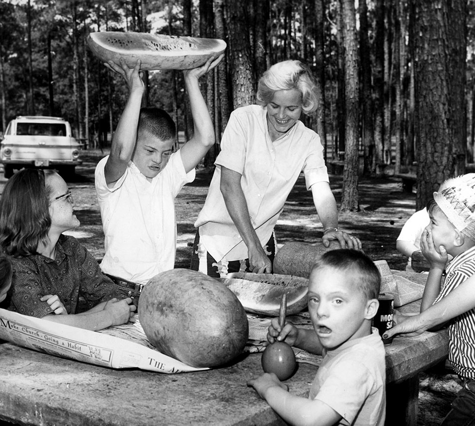 Black and white photo of woman and children sitting on a bench in a park. Boy with Down syndrome is holding a halved watermelon over his head.