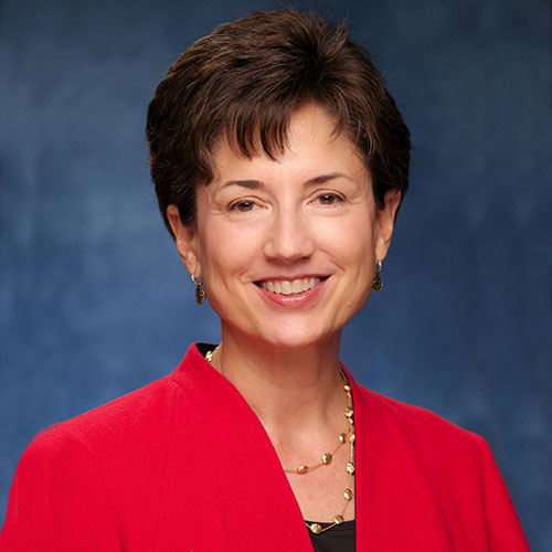Head shot of a woman with short brown hair and a red blazer in front of a blue background